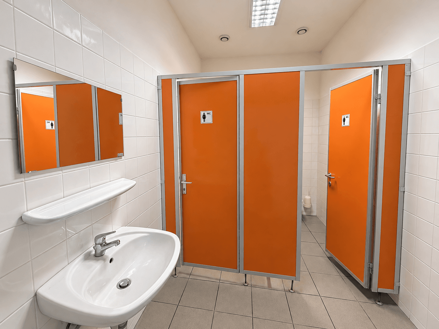 Öffentliche Toilettenanlage mit weißen Fliesen, orangefarbenen Kabinentüren und einem Waschbecken.