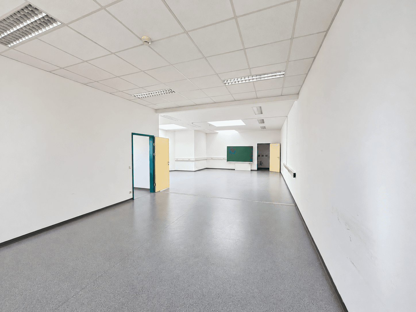 Langer, leerer Flur oder offener Raum mit mehreren Türen und einer Tafel im Hintergrund.