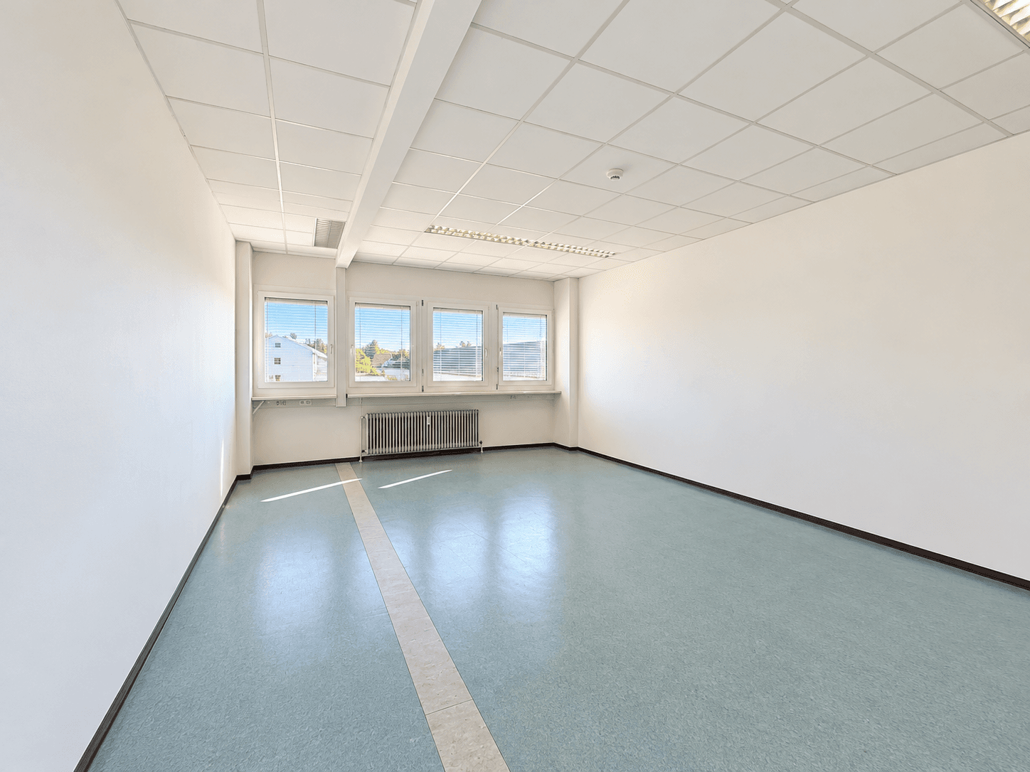 Leerer Büroraum mit mehreren Fenstern, Heizkörpern und einer Deckenkonstruktion mit Leuchten.