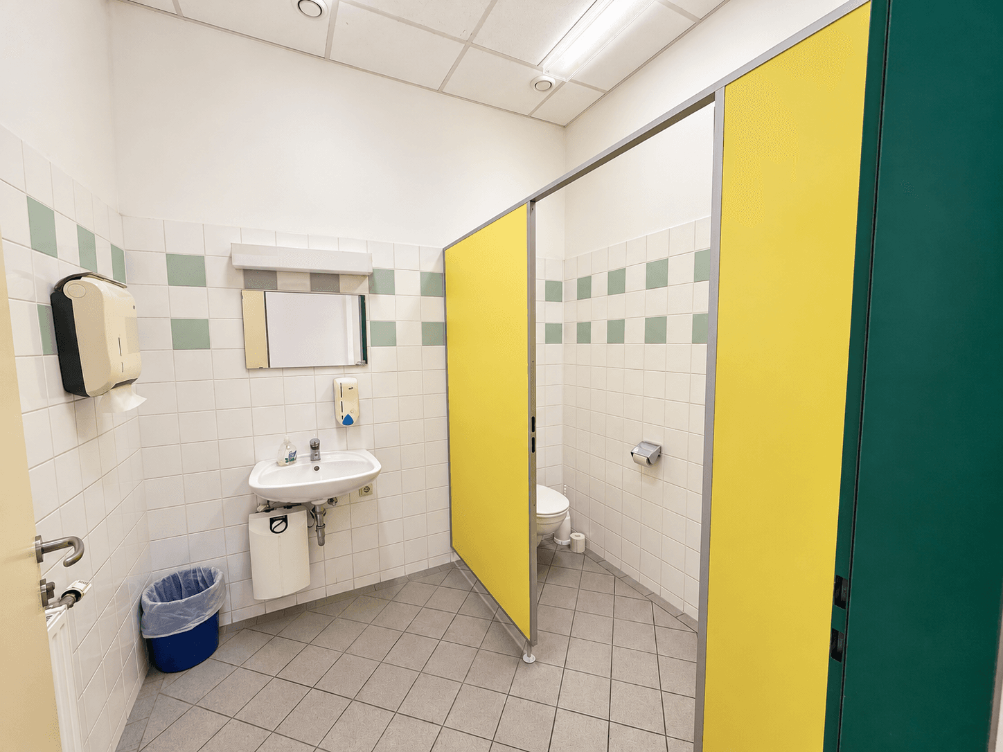 Öffentliche Toilette mit mehreren Kabinen, Waschbecken und gefliesten Wänden und Böden.