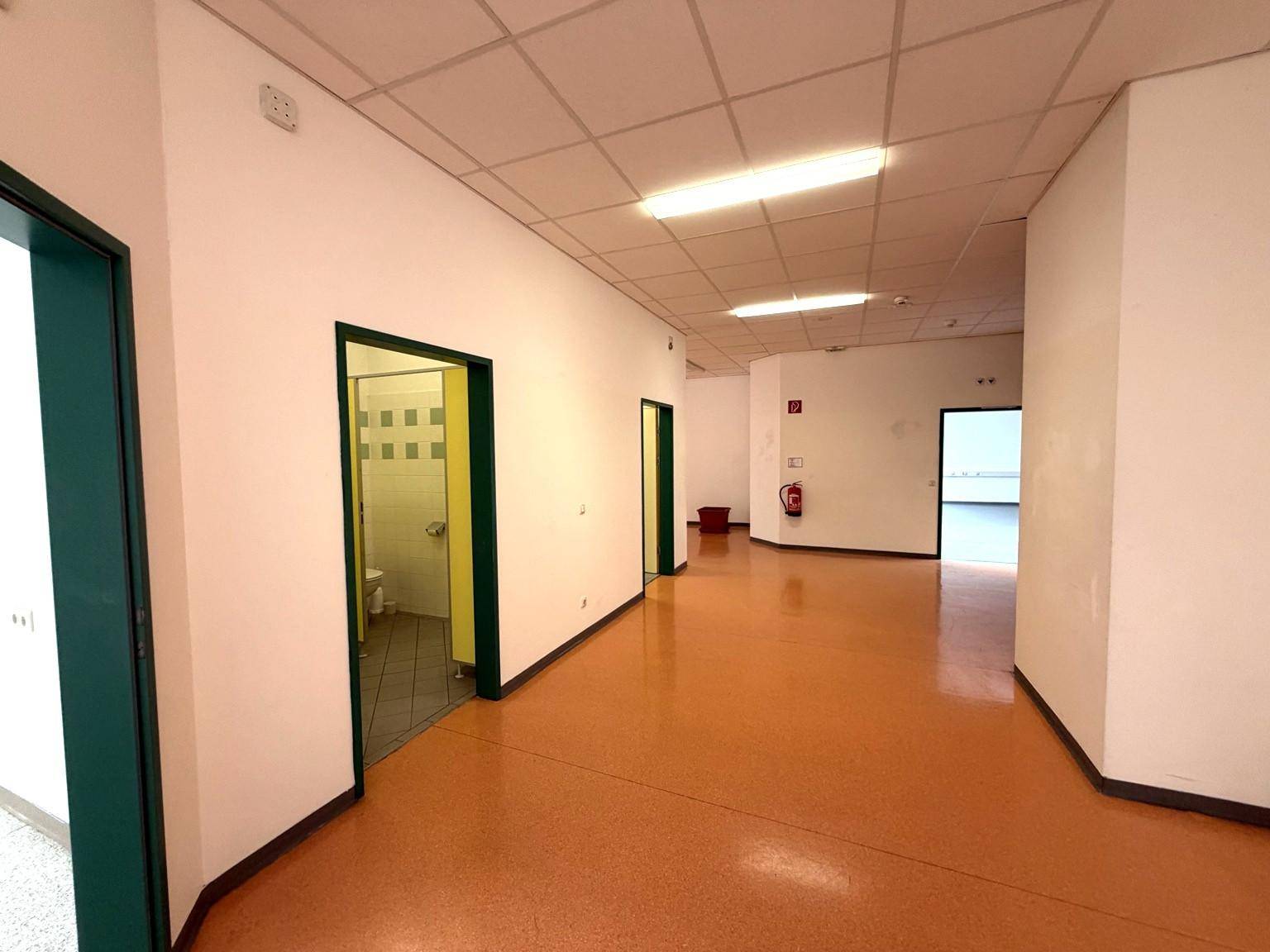Langer Flur mit mehreren Türen, einem Feuerlöscher und einem orangefarbenen Bodenbelag.