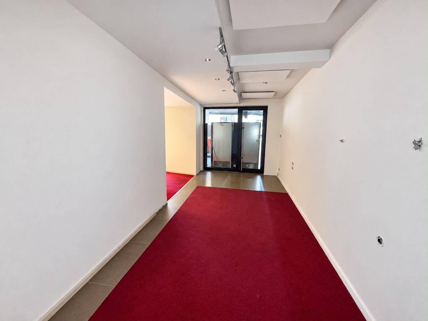 Langer, schmaler Geschäftsraum mit rotem Teppich, weißen Wänden und Glastüren am Ende des Ganges.