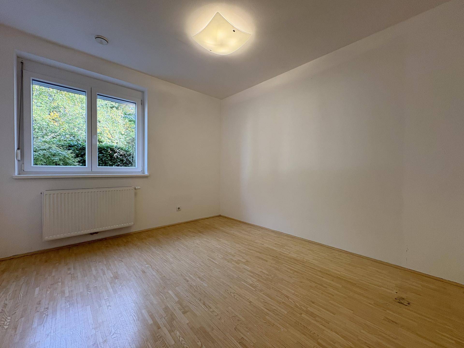 Helles Schlafzimmer mit Holzboden und Fenster mit Blick ins Grüne, bereit zur individuellen Gestaltung.