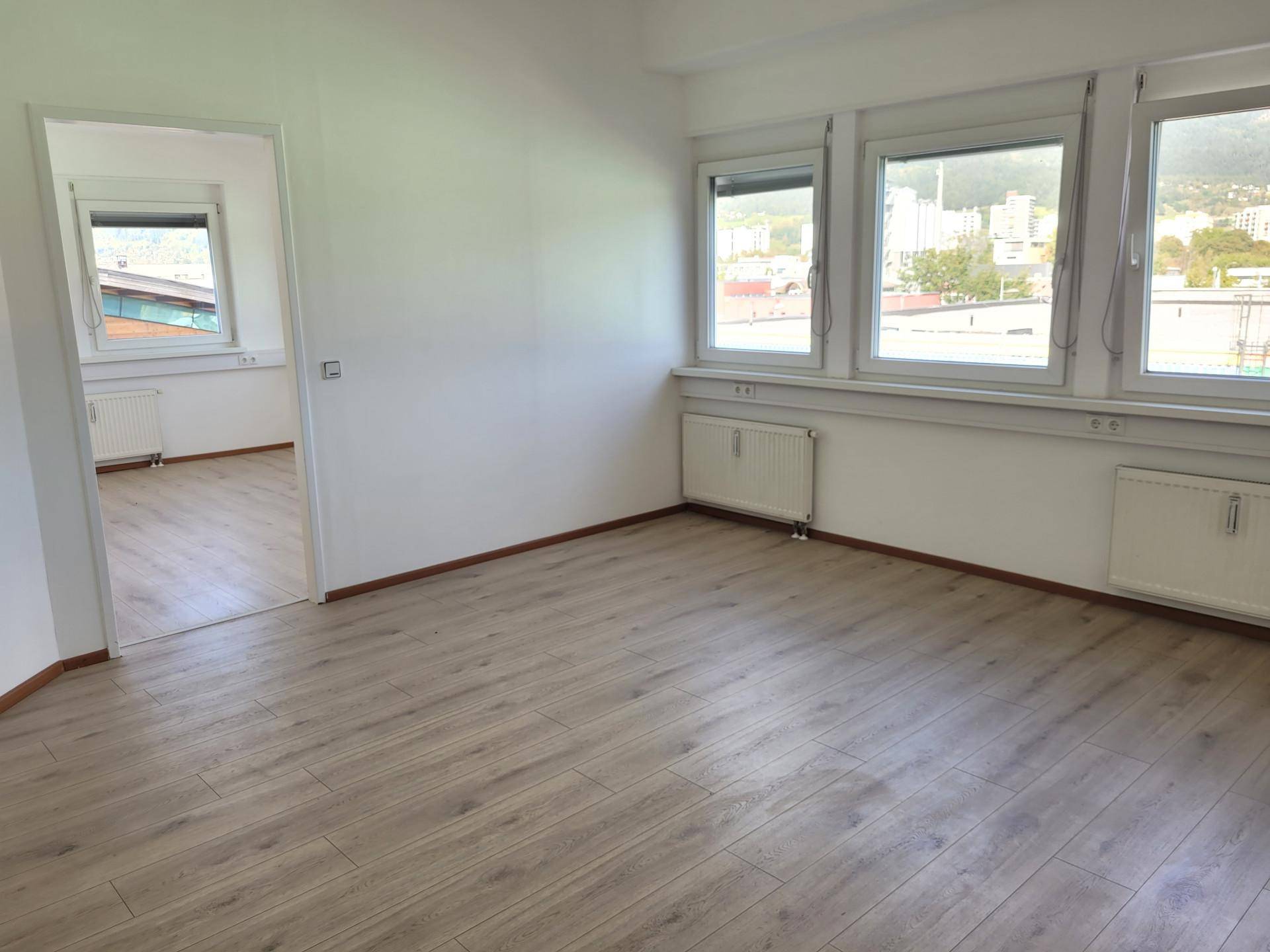 Leerer Büroraum mit Laminatboden, großen Fenstern und Heizkörpern unter den Fensterbänken.