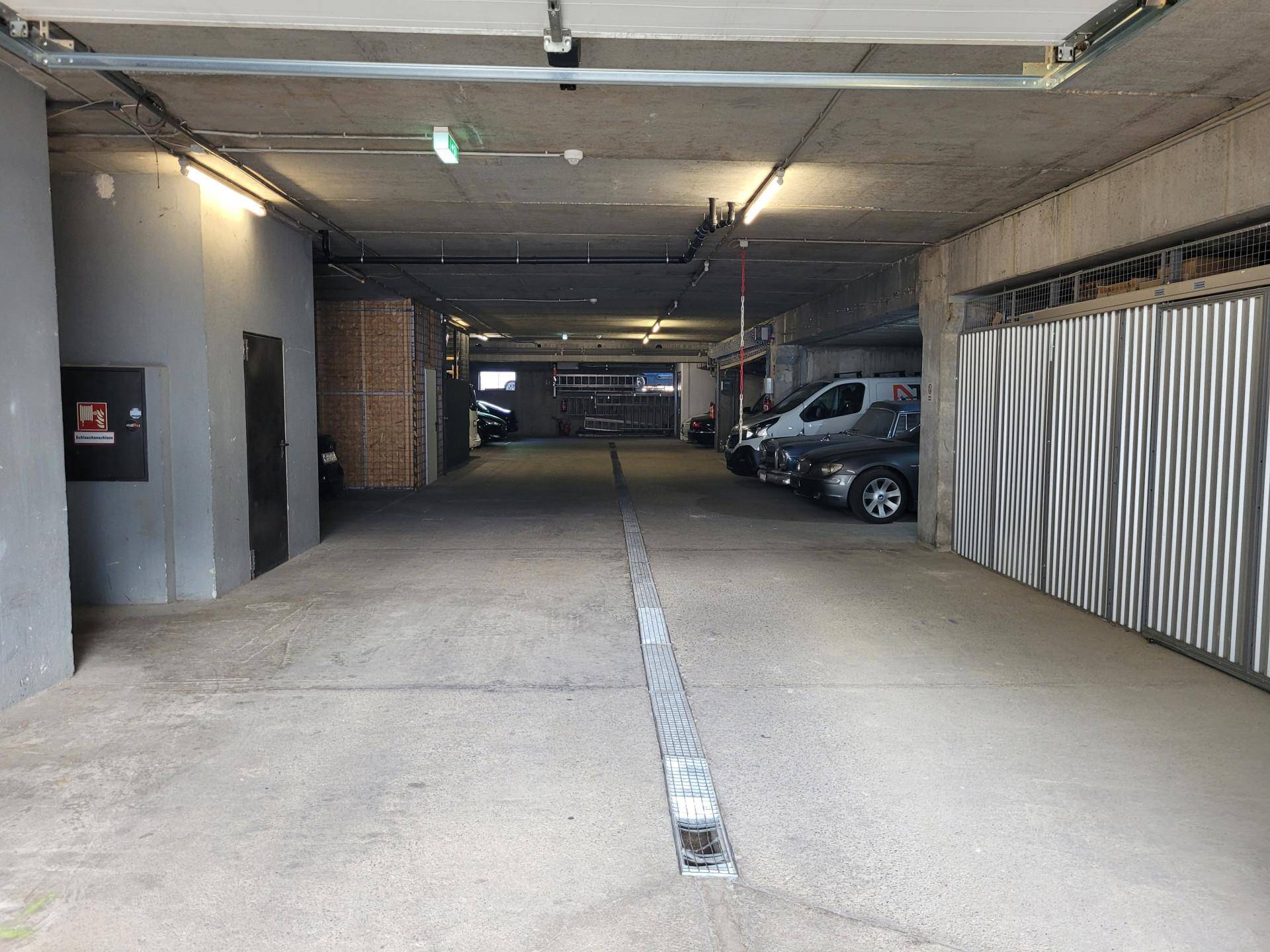Tiefgarage mit Betonboden, Deckenbeleuchtung und mehreren geparkten Fahrzeugen auf der rechten Seite.