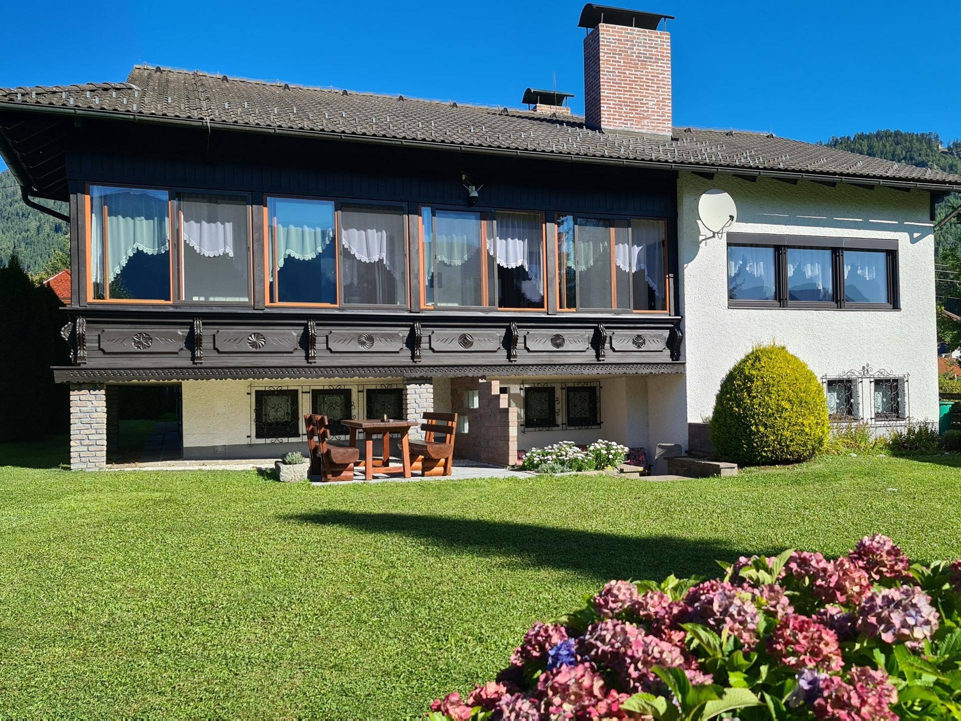 Traditionelles Haus mit Holzbalkon und gepflegtem Gartenbereich im Vordergrund unter blauem Himmel.