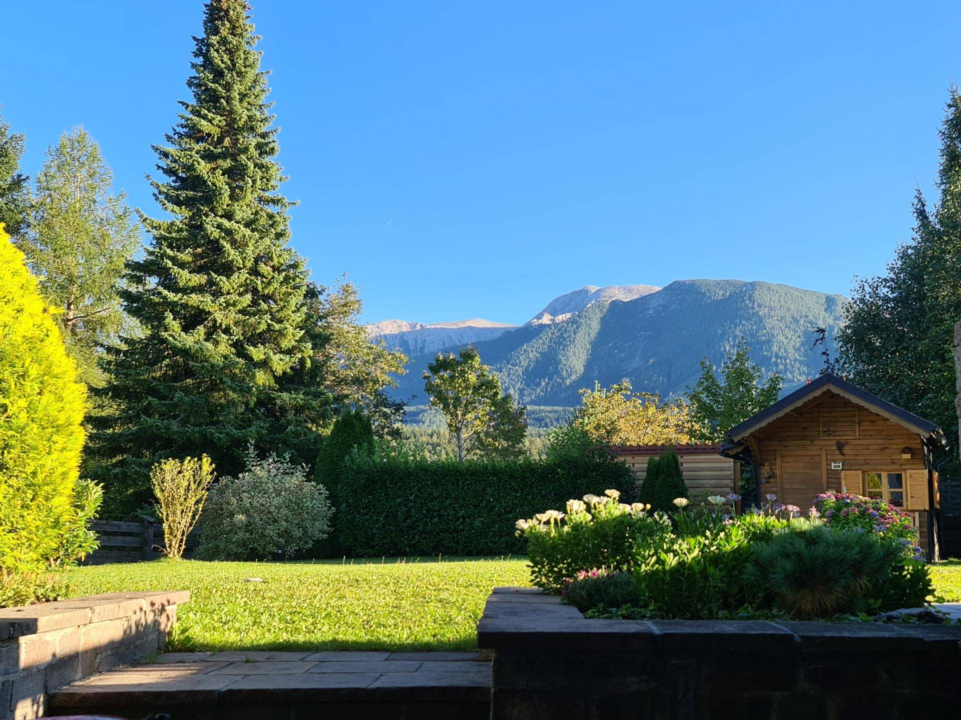 Gepflegter Garten mit Blumenbeeten, hohen Bäumen und Bergblick im Hintergrund an einem sonnigen Tag.