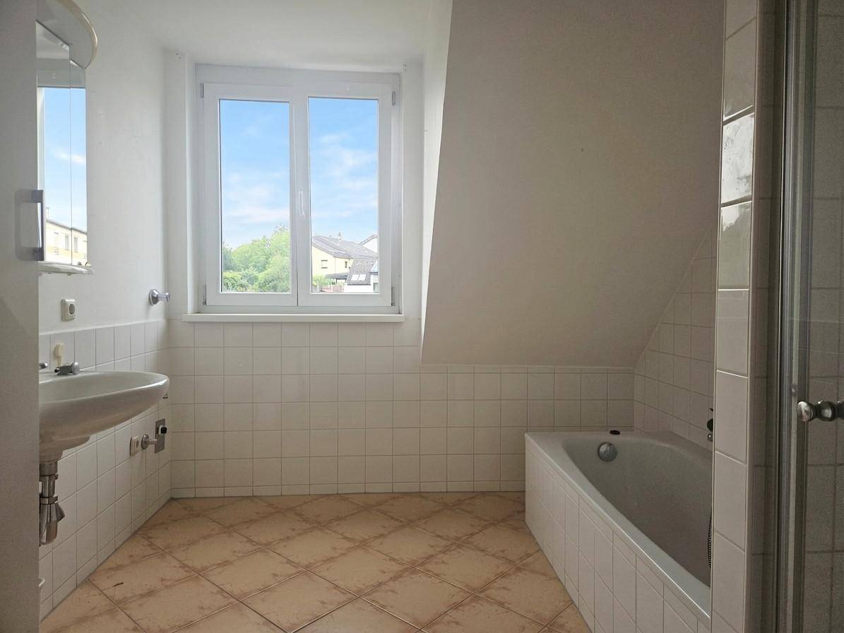 Helles Badezimmer im Dachgeschoss mit Badewanne, Dusche, Waschbecken und Fenster, ausgestattet mit Fliesenboden.