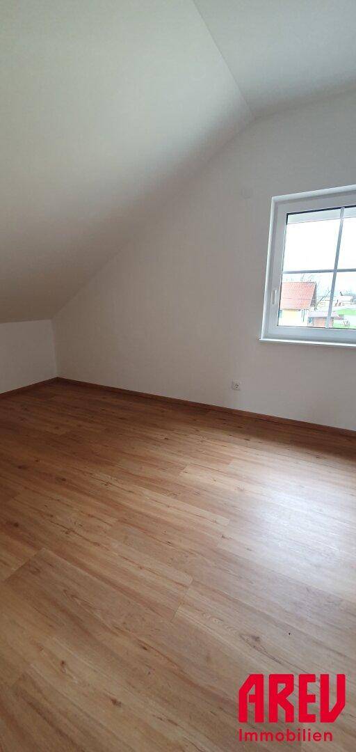 Helles Schlafzimmer im Dachgeschoss mit Holzboden und einem Fenster mit Blick ins Grüne.