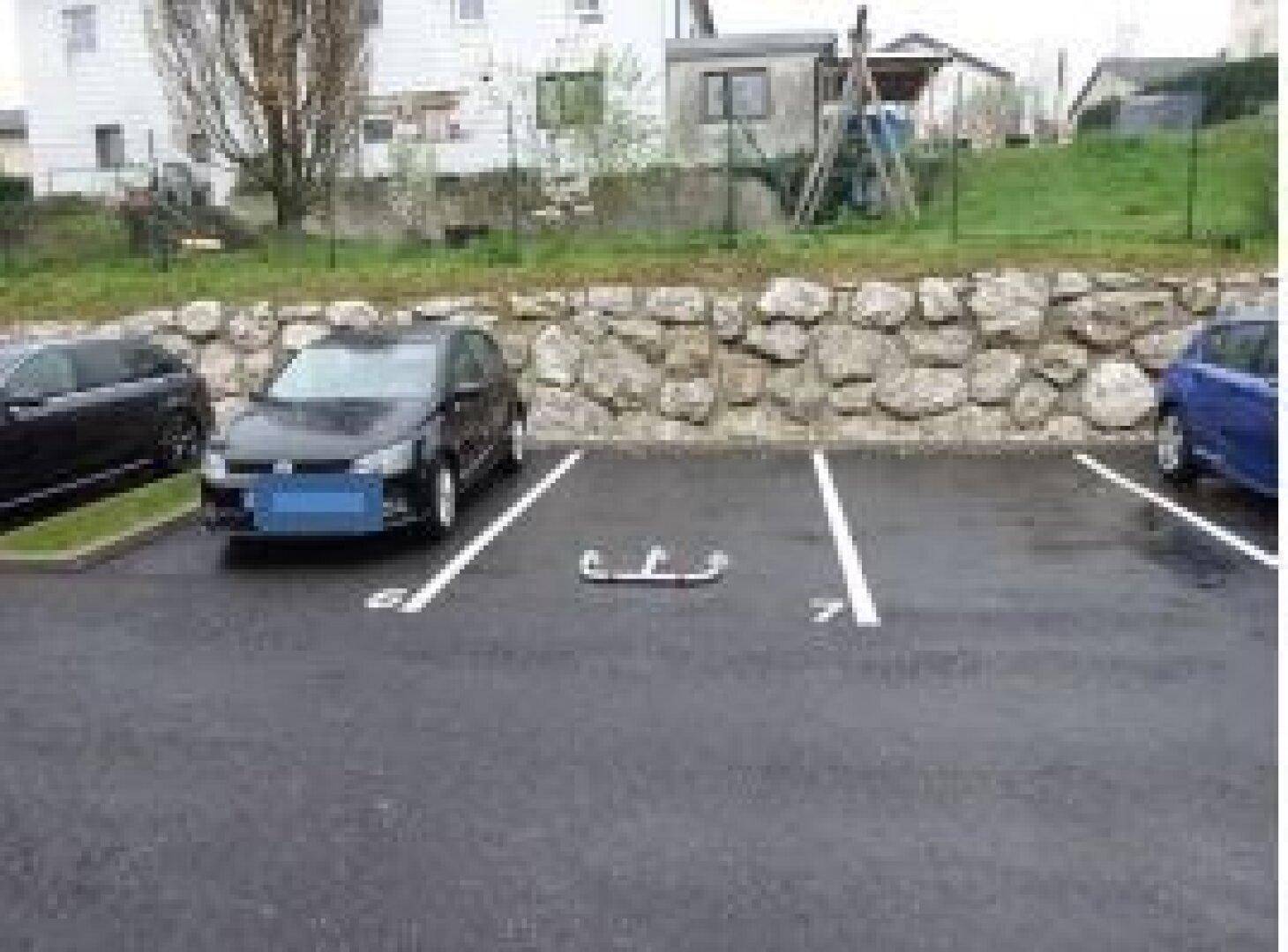 Mehrere Außenstellplätze auf einem asphaltierten Parkplatz vor einer Steinmauer und Grünfläche.