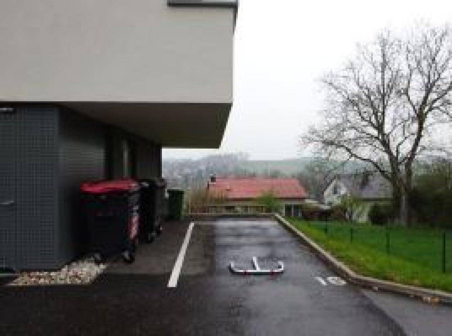 Außenansicht des Gebäudes mit Mülltonnen und einem Stellplatz, umgeben von ländlicher Landschaft.