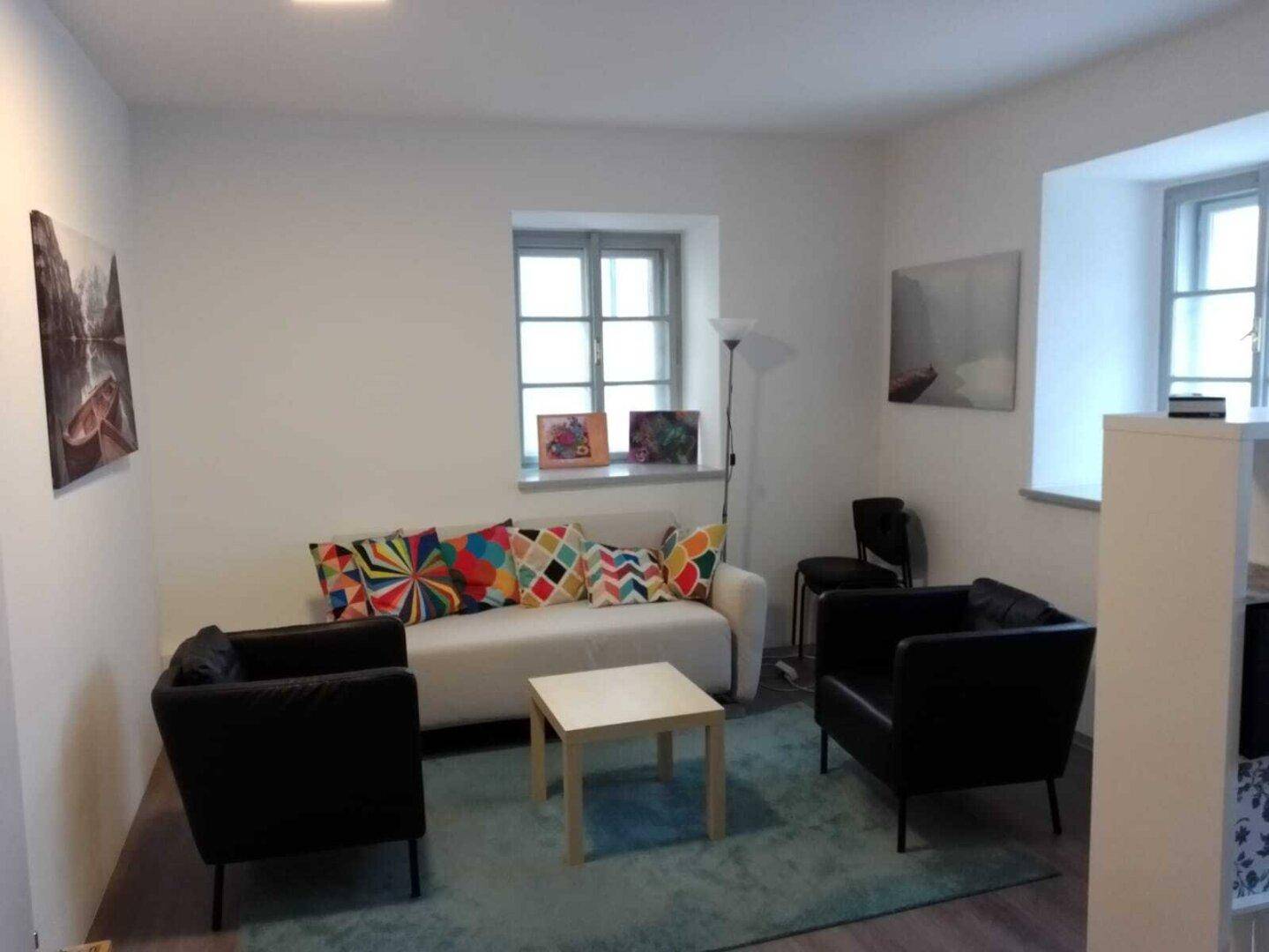 Gemütlicher Wartebereich mit einem hellen Sofa, bunten Kissen, zwei Sesseln und zwei Fenstern.