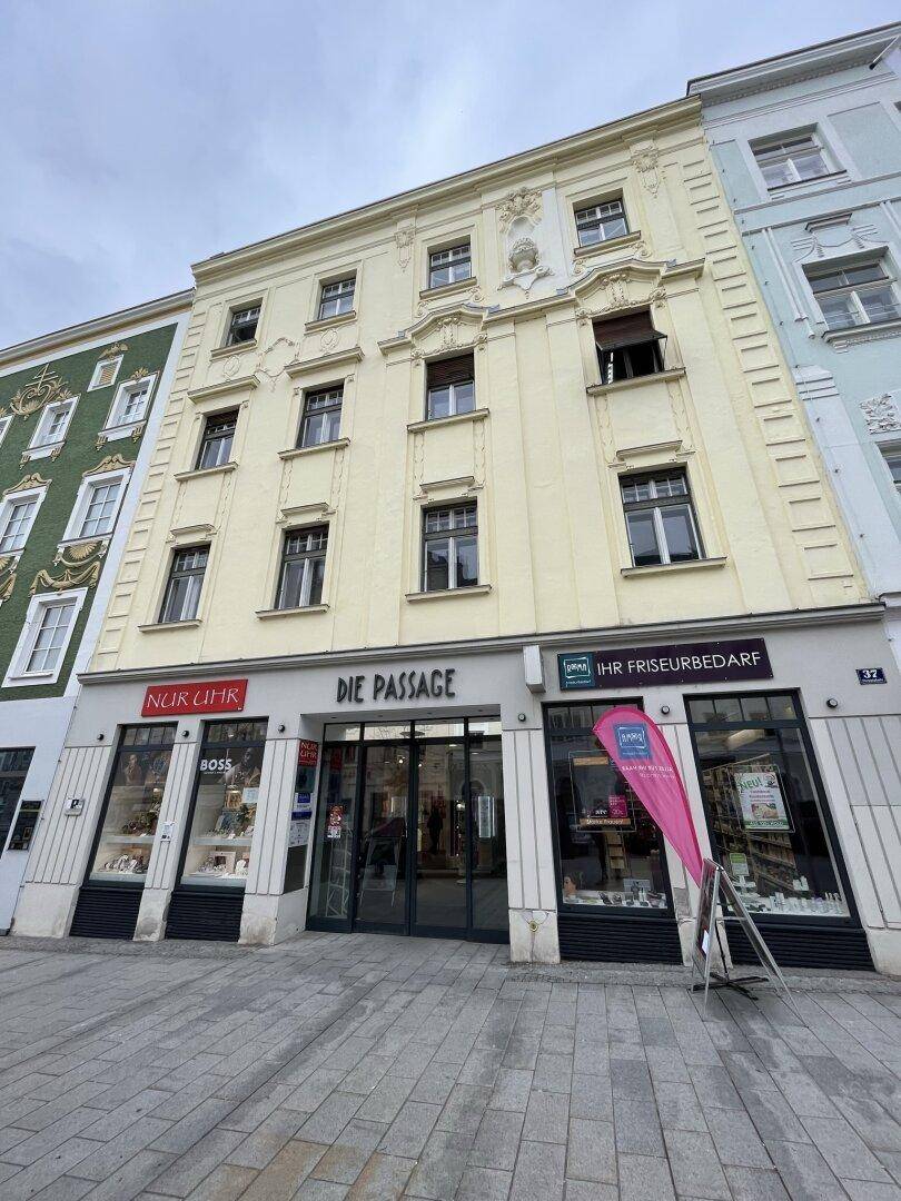 Historische Gebäudefassade mit Geschäften im Erdgeschoss und dekorativen Fenstern in der oberen Etage, an einer belebten Straße gelegen.