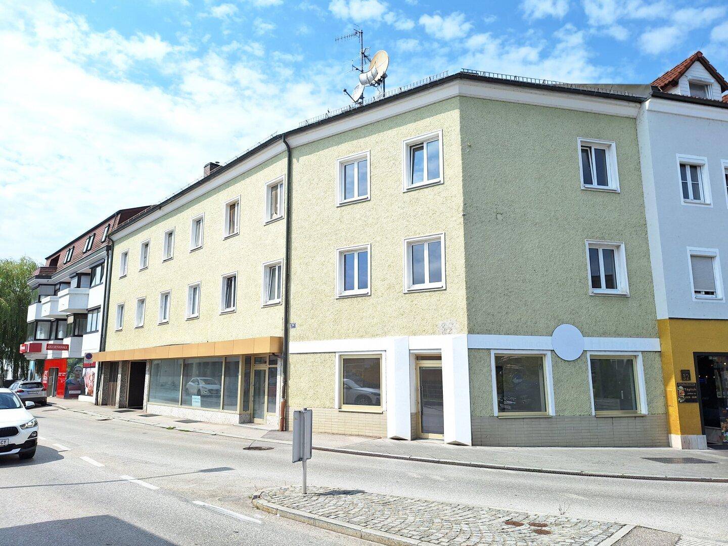 Mehrgeschossiges Gebäude mit hellgrüner Fassade an einer Straße, mit Geschäften im Erdgeschoss und Wohnungen darüber.