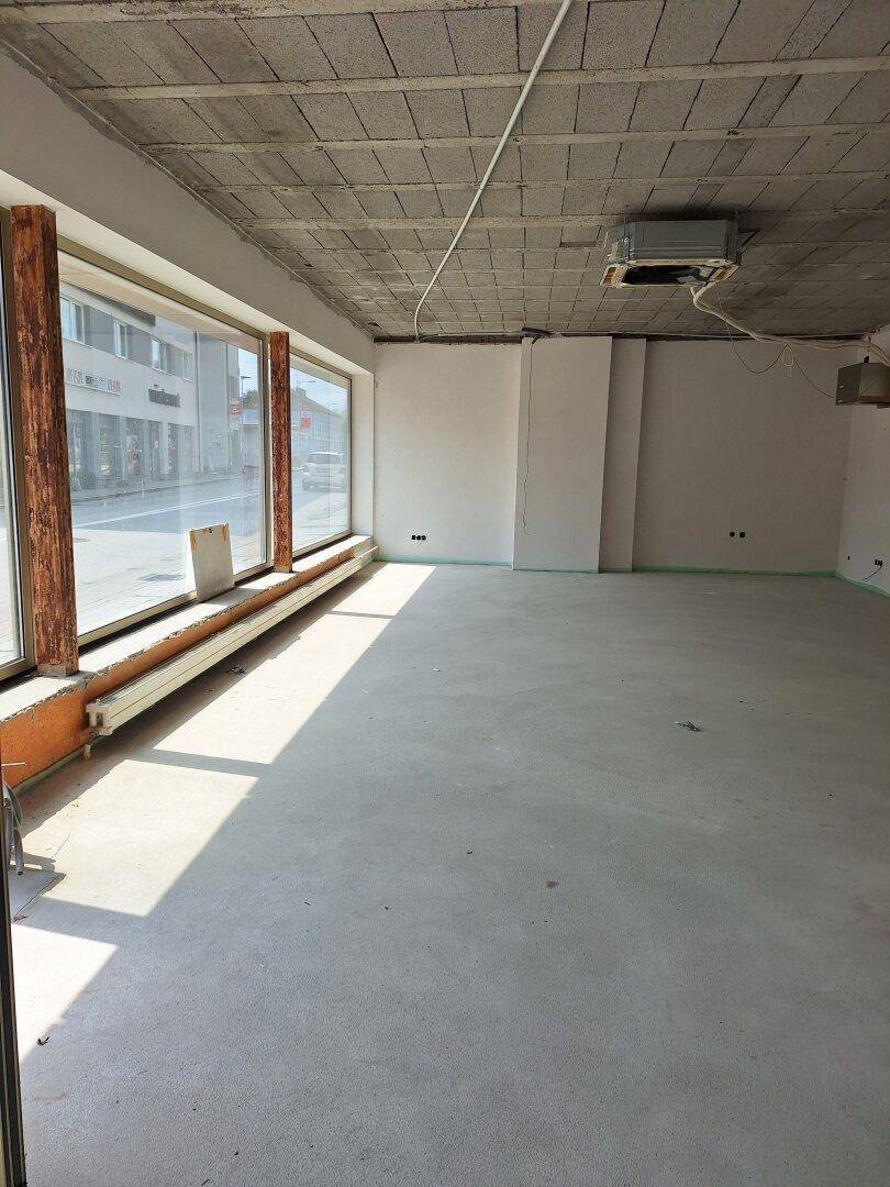 Großer, leerer Raum im Rohbau mit Betonboden, unverputzter Decke und großen Fenstern zur Straße.