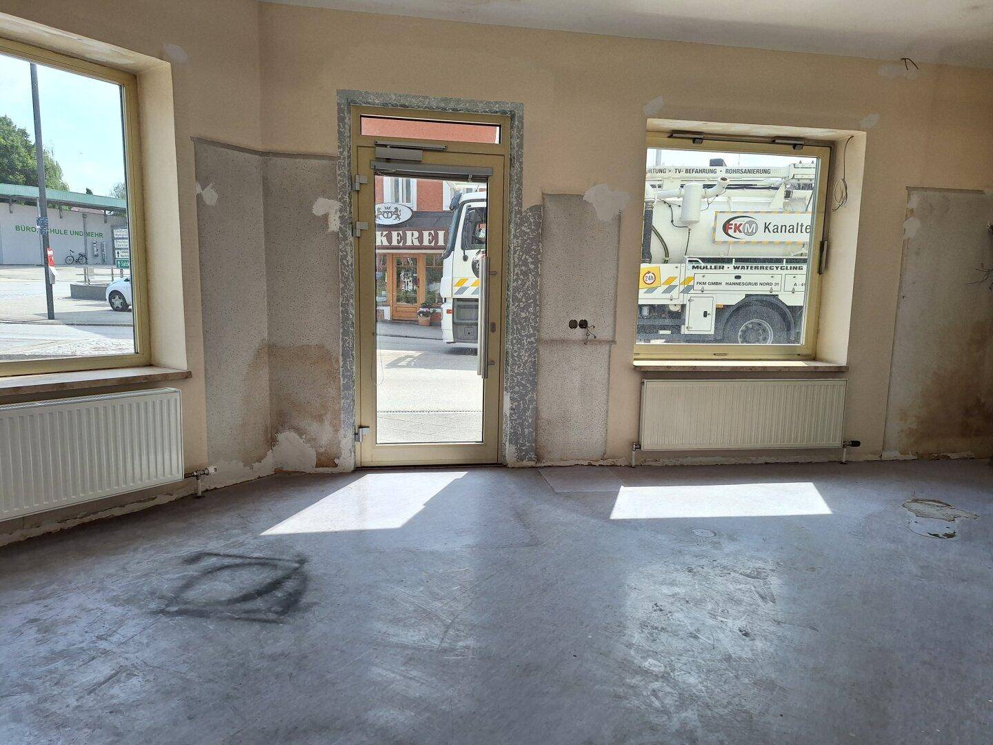 Renovierungsbedürftiger Raum mit großen Fenstern, Heizkörpern und einer Eingangstür zur Straße.