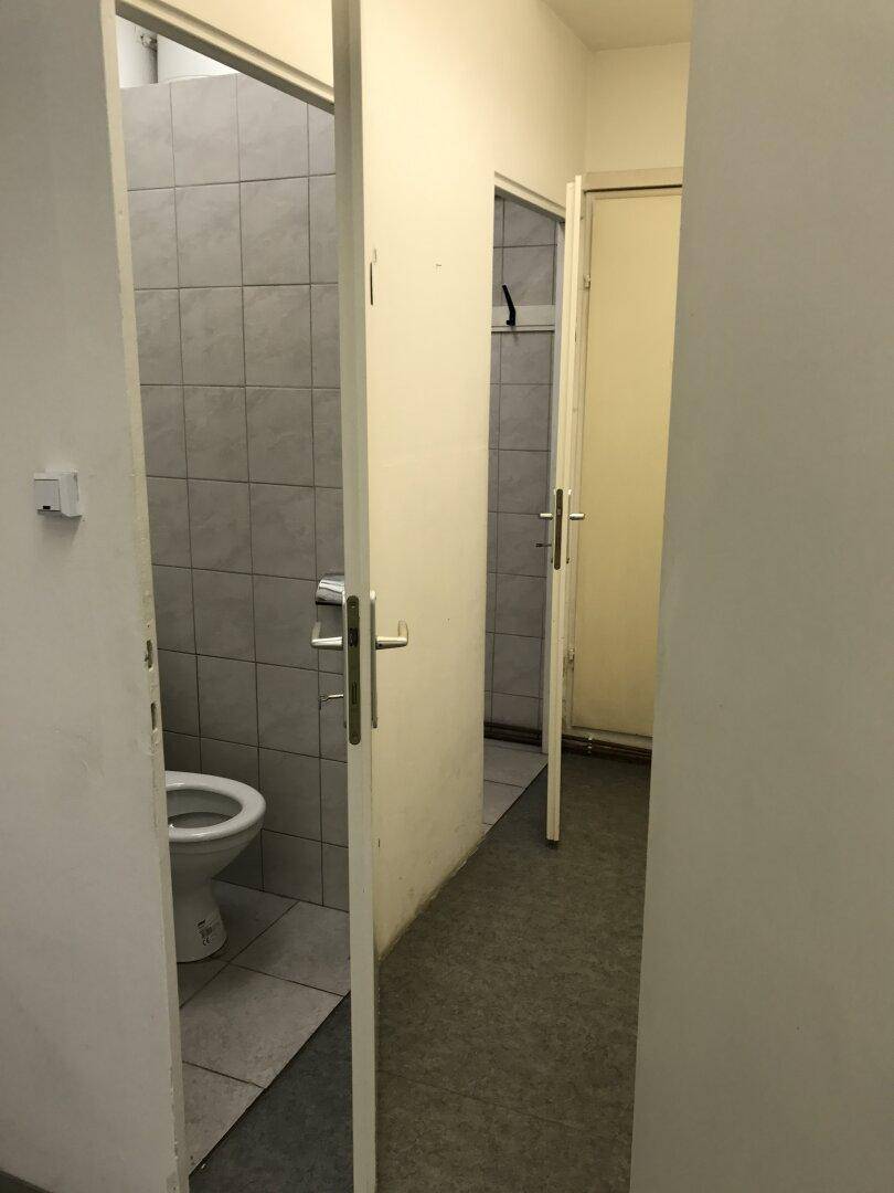 Schmaler Flur führt zu zwei Toilettenkabinen mit grauen Fliesen und einfachen Sanitäranlagen.