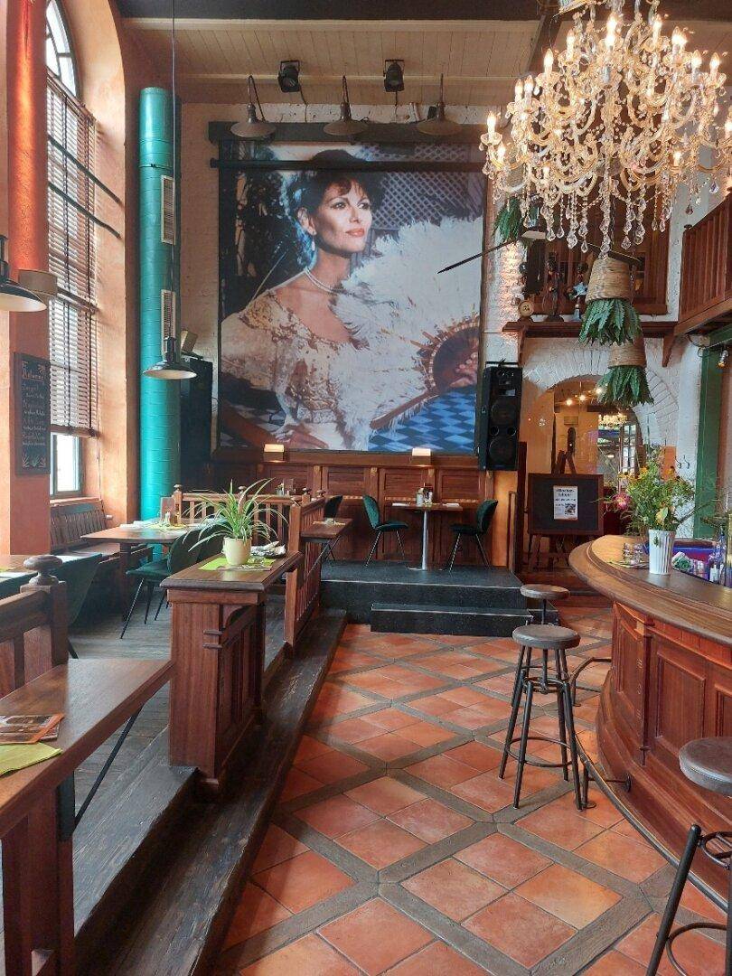 Geräumiger Restaurantbereich mit hohen Decken, großen Bogenfenstern, einem Kronleuchter und einer Bar.