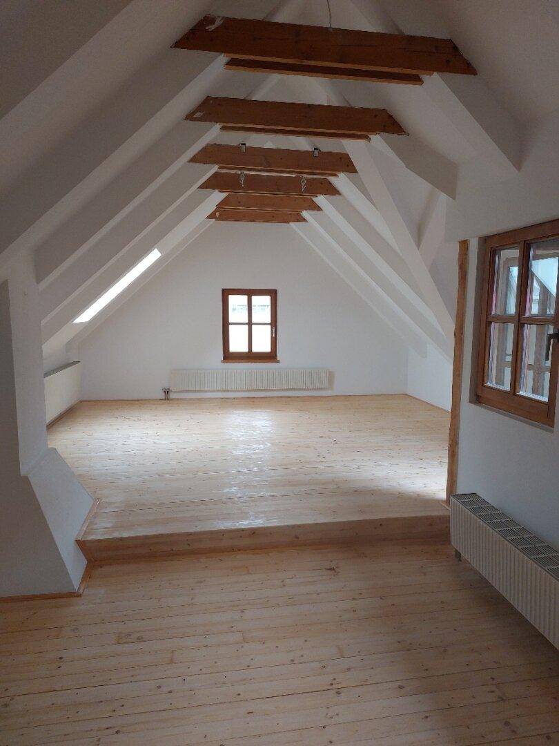 Großzügiger Dachbodenraum mit Holzdielenboden, sichtbaren Balken und einem Fenster.