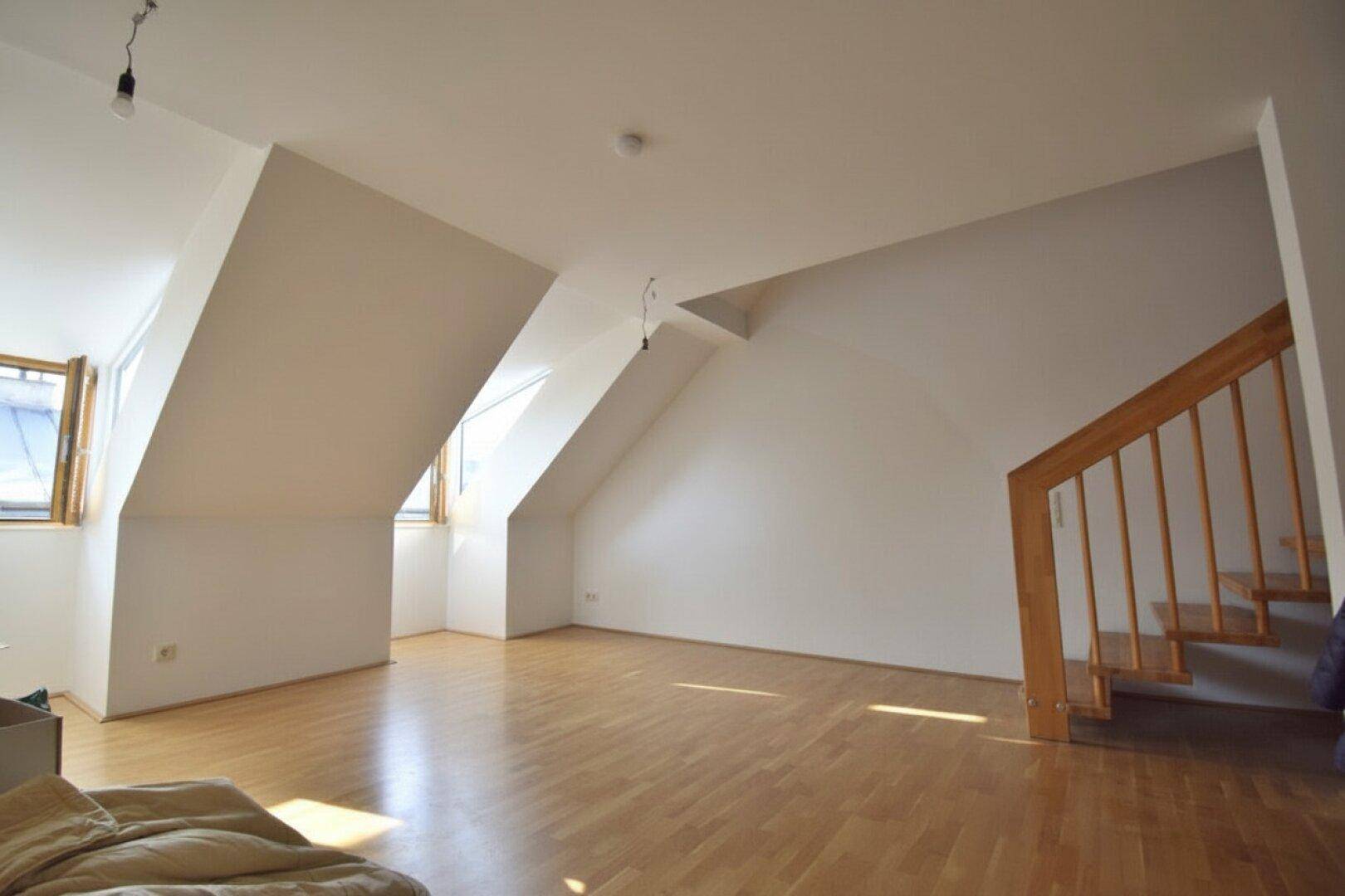 Geräumiger Raum mit Dachschrägen, Holzboden und einer Holztreppe.