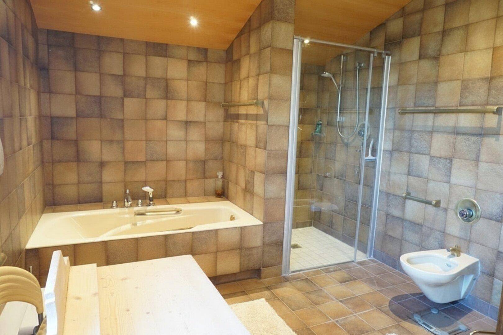 Geräumiges Badezimmer mit Badewanne, Duschkabine und Fliesen in warmen Brauntönen.