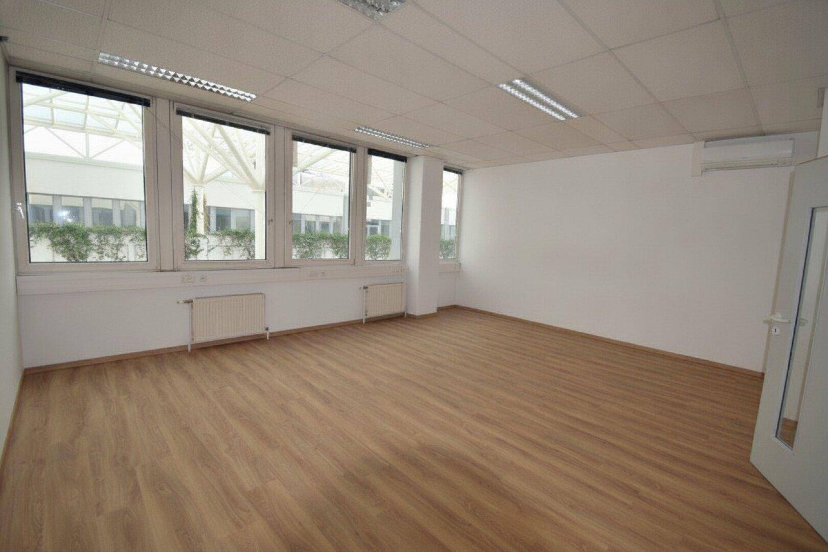 Leerer Büroraum mit großen Fenstern, Laminatboden und Klimaanlage, bereit zur individuellen Gestaltung.