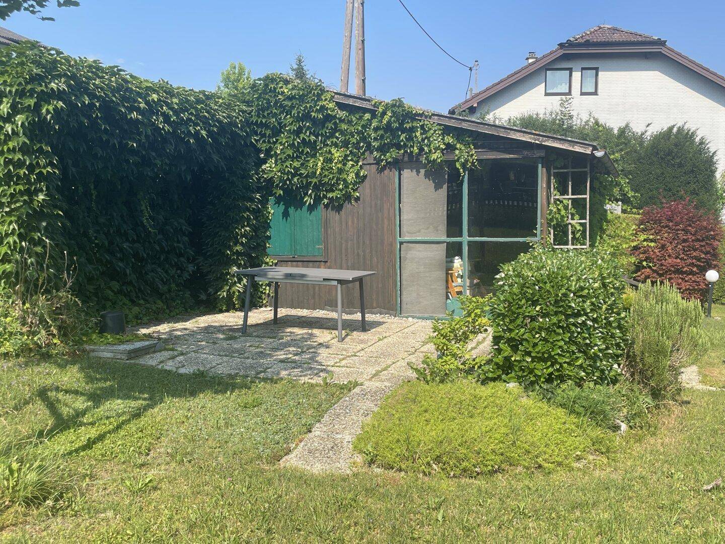 Gemütliche Terrasse vor einem Holzschuppen, umgeben von üppigem Grün und Efeu.