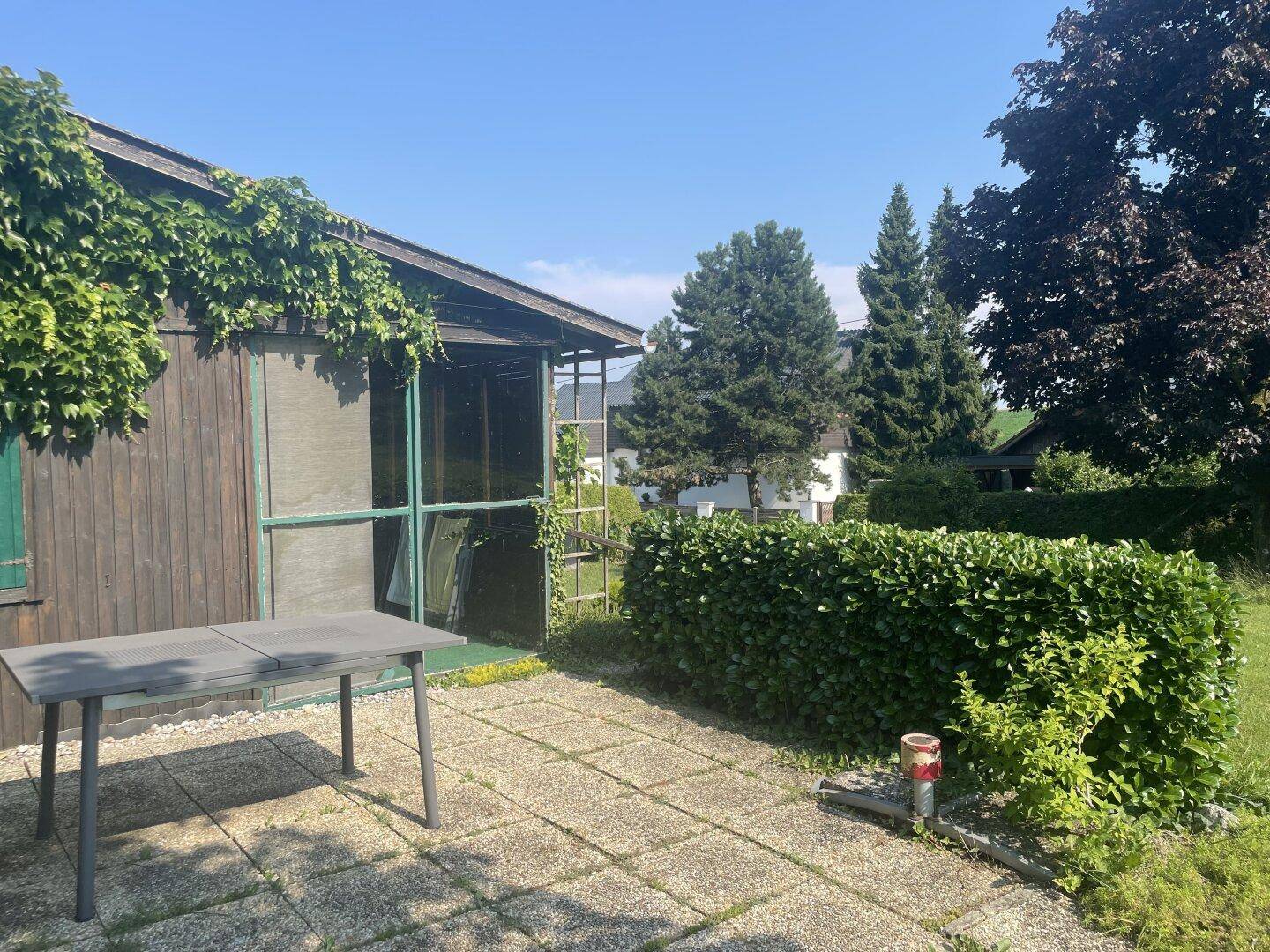 Sonniger Terrassenbereich mit Pflastersteinen vor einem Holzgebäude, umgeben von Hecken und Bäumen.