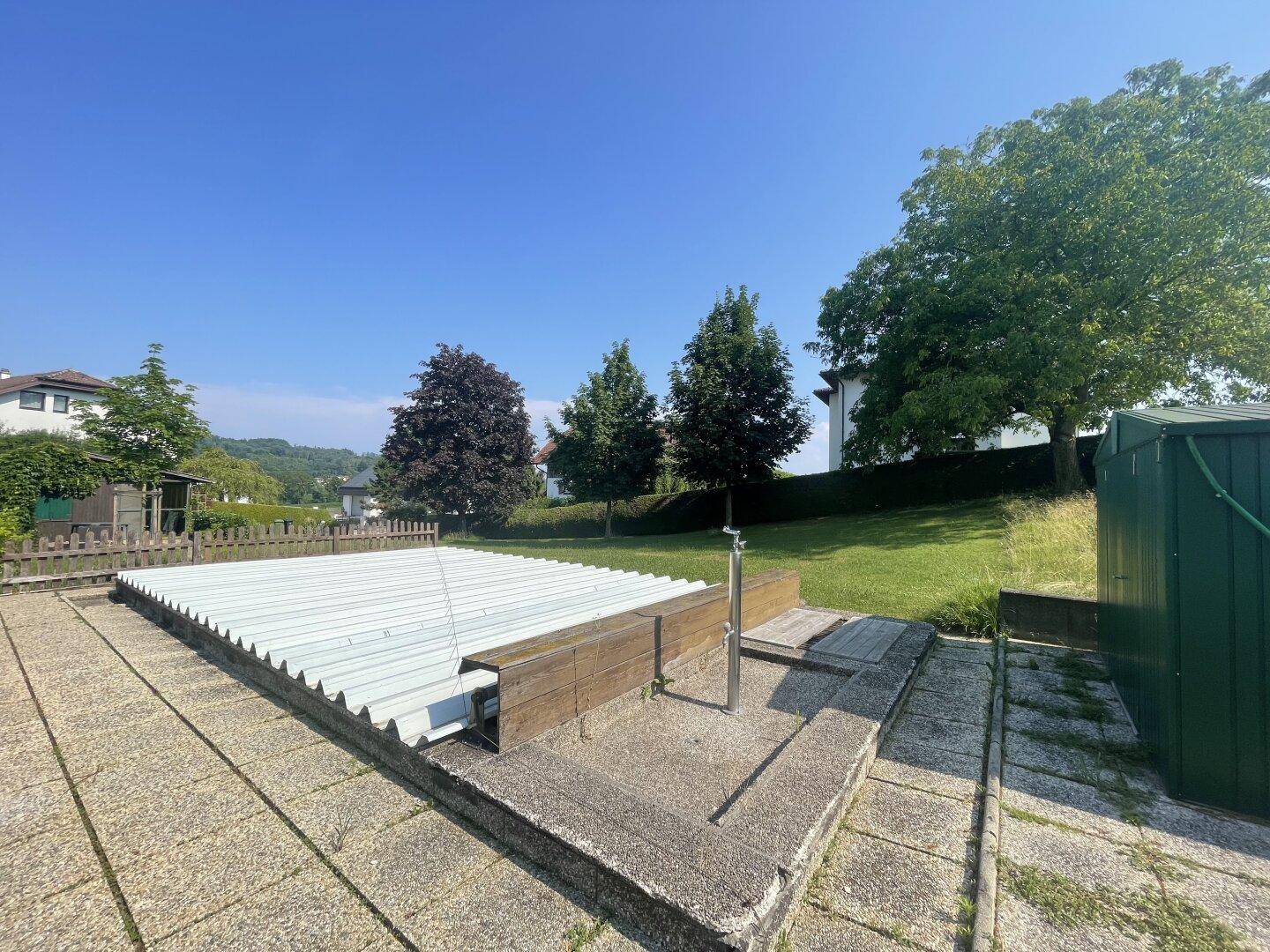 Abgedeckter Swimmingpool mit gepflastertem Bereich und Außendusche in einem sonnigen Garten.