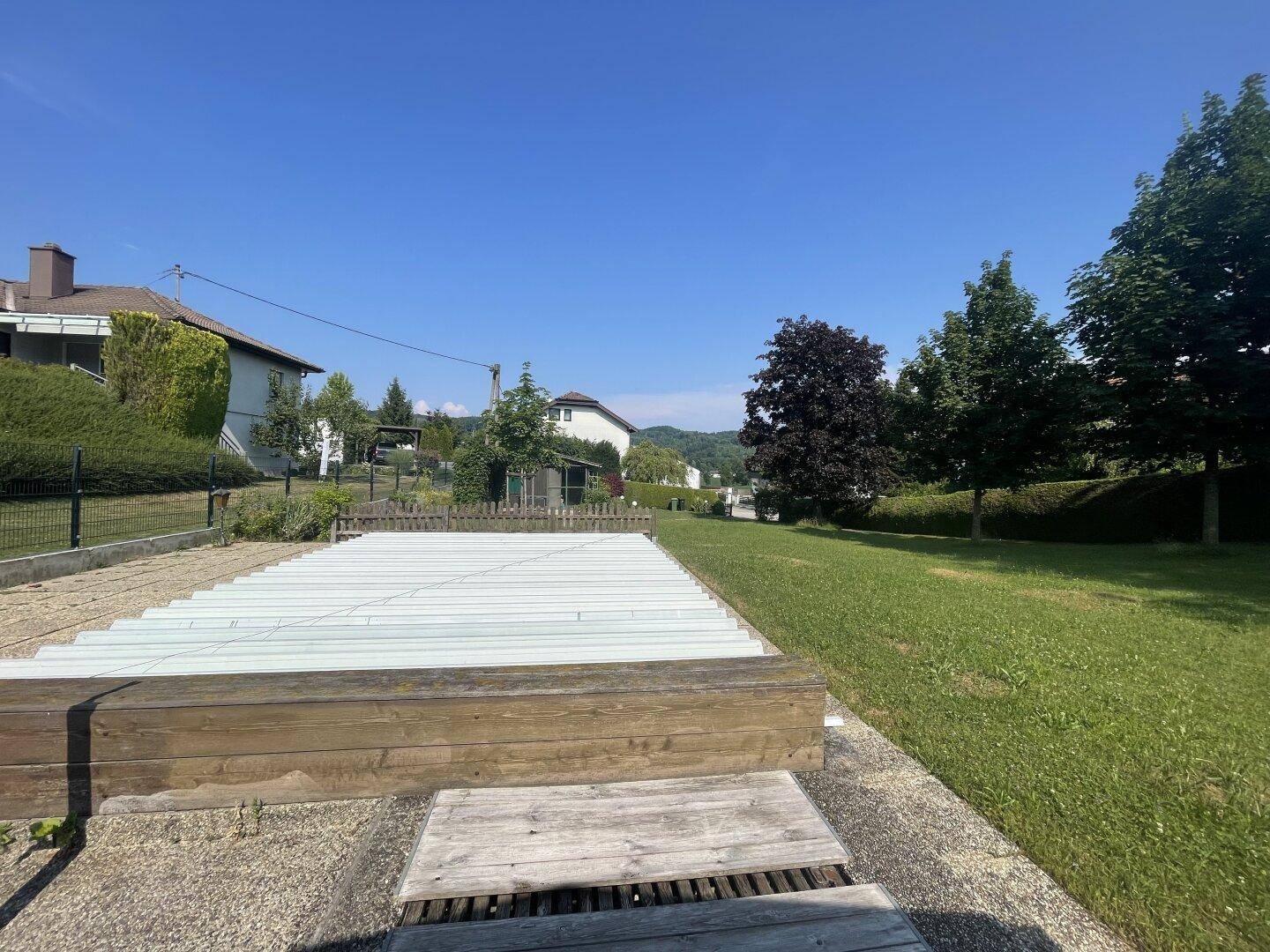 Blick auf den abgedeckten Poolbereich und die weitläufige Rasenfläche des Gartens an einem sonnigen Tag.