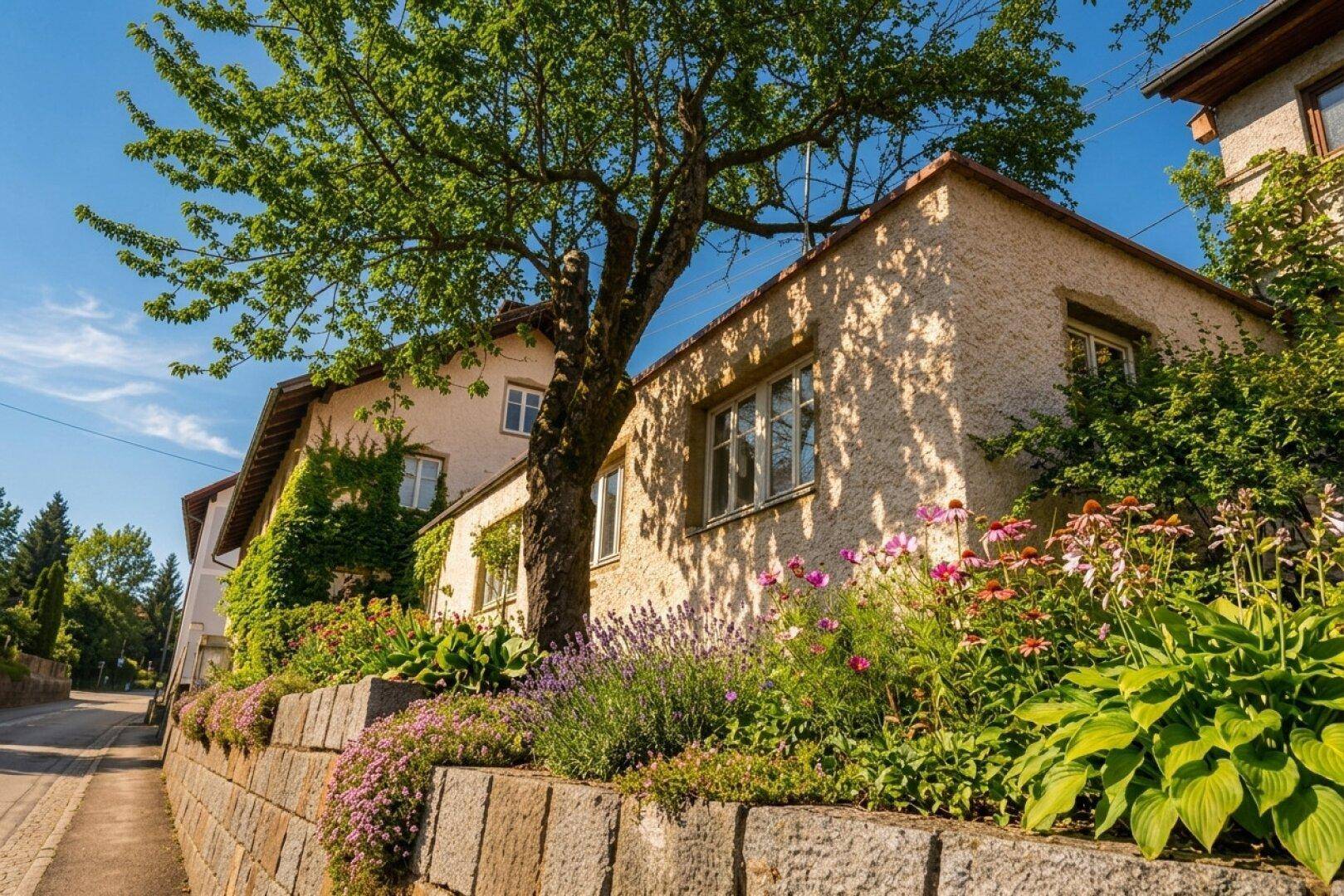 Hausfassade mit blühenden Blumenbeeten, einem großen Baum und einer Steinmauer unter blauem Himmel.