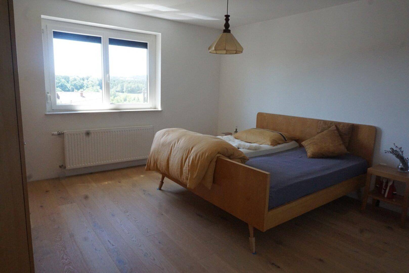 Helles Schlafzimmer mit Holzbett, Parkettboden und Fenster mit Weitblick ins Grüne.