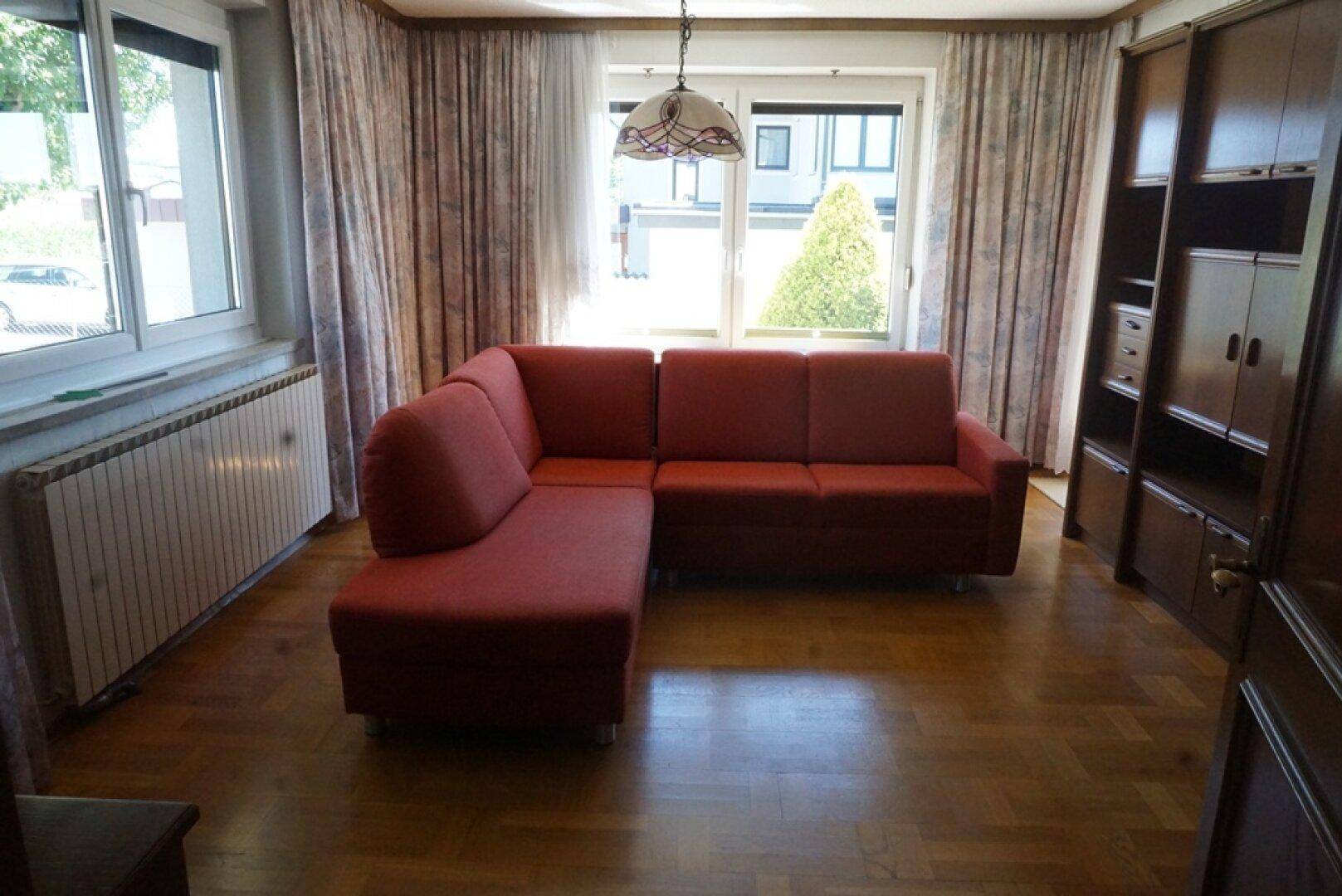 Gemütliches Wohnzimmer mit rotem Ecksofa, Parkettboden und Blick ins Freie.