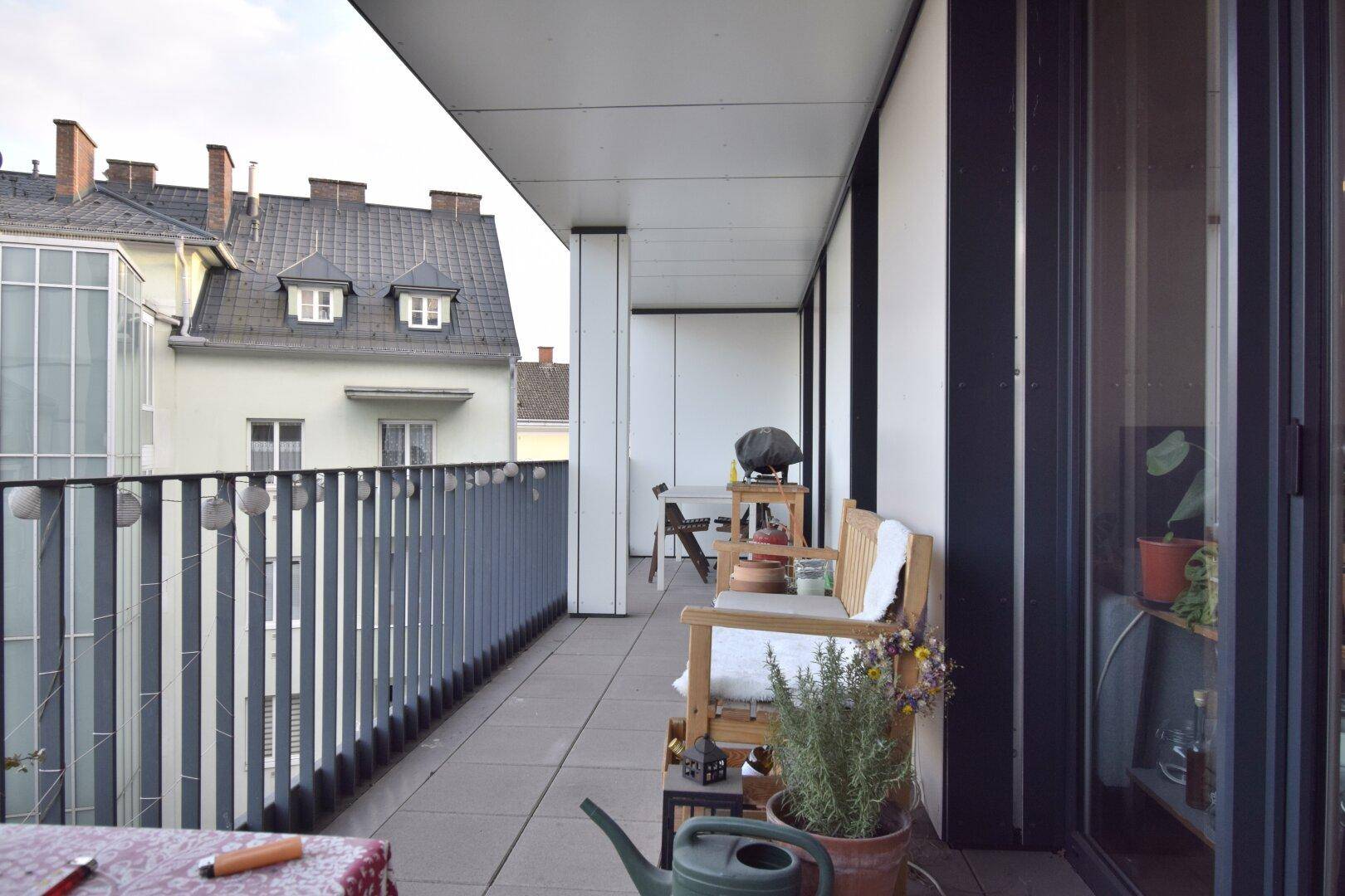 Langer Balkon mit Fliesenboden, Sitzgelegenheiten und Blick auf die umliegenden Gebäude.