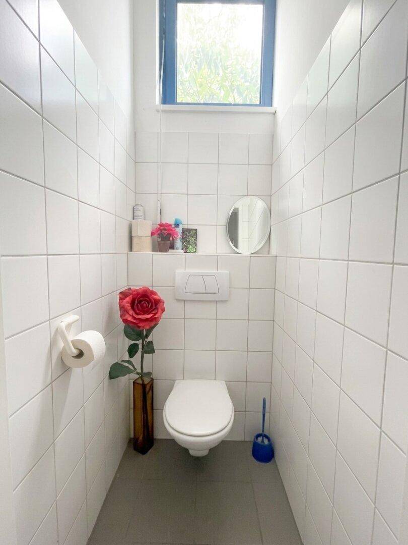 Kleiner Toilettenraum mit weißen Fliesen, wandhängendem WC und einem Fenster für Belüftung und Tageslicht.