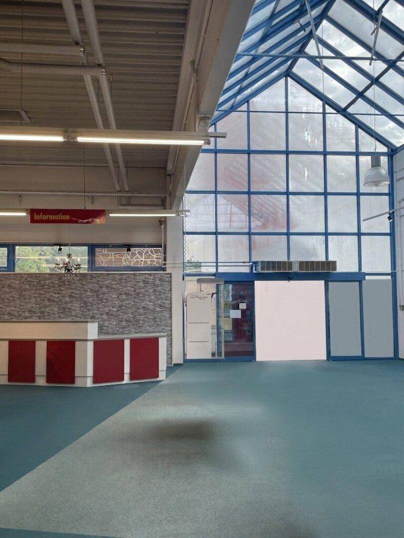 Empfangsbereich eines Geschäftsraums mit moderner Theke, Teppichboden und einer hohen Glasfassade, die viel Licht hereinlässt.