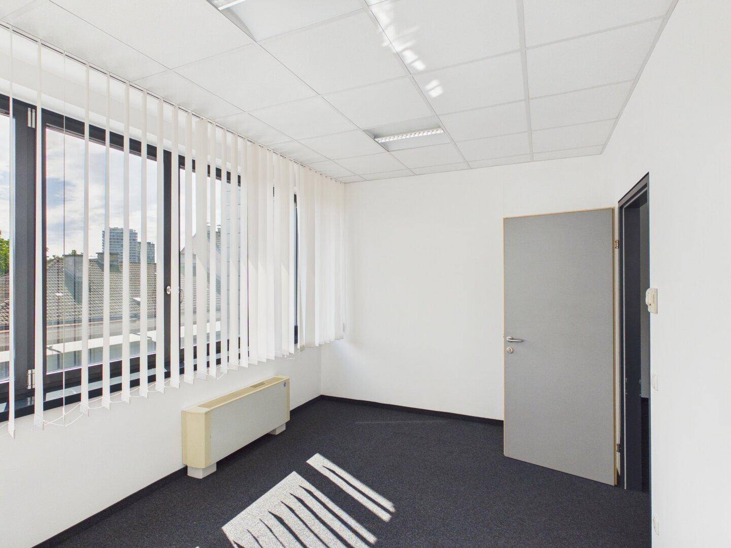 Kleines, helles Büro mit Teppichboden, vertikalen Jalousien und Blick auf die Stadt.