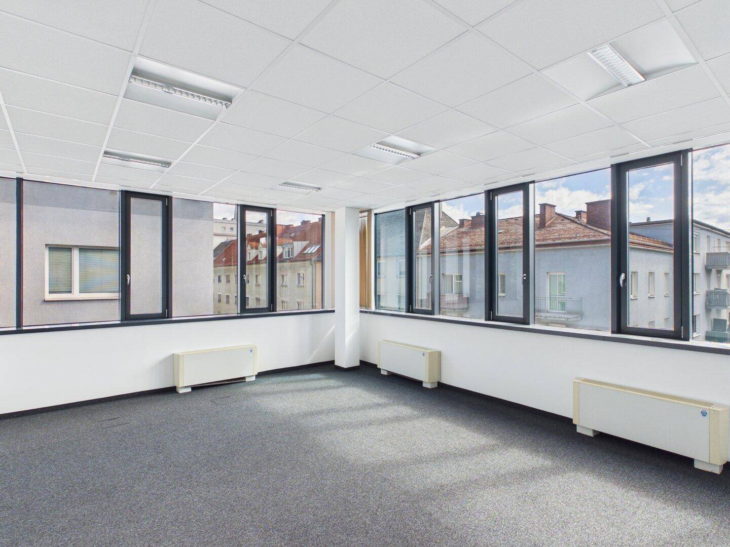 Helles Büro mit Teppichboden, großen Fenstern und Blick auf die umliegenden Gebäude.