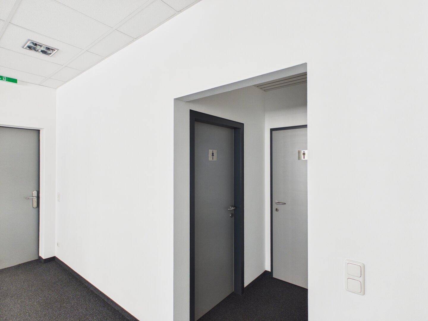 Flur mit Teppichboden und Türen zu den Toilettenbereichen in einem Bürogebäude.