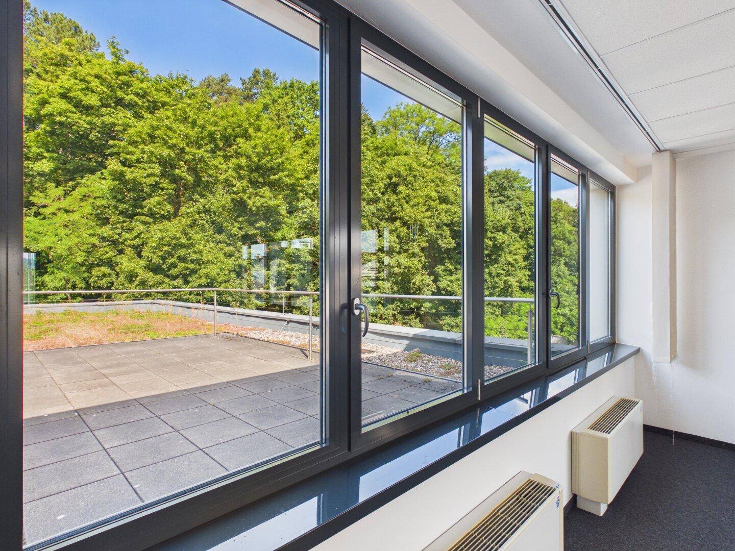 Büro mit großen Fenstern, die eine Dachterrasse und grüne Bäume zeigen.