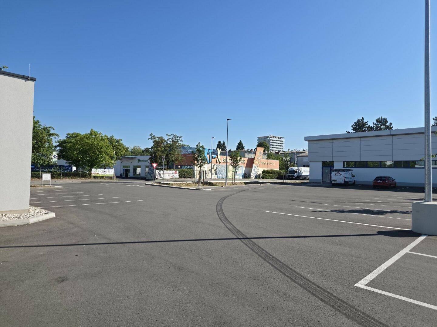Großer, leerer Parkplatz vor Gewerbegebäuden unter strahlend blauem Himmel, bereit für Kunden.