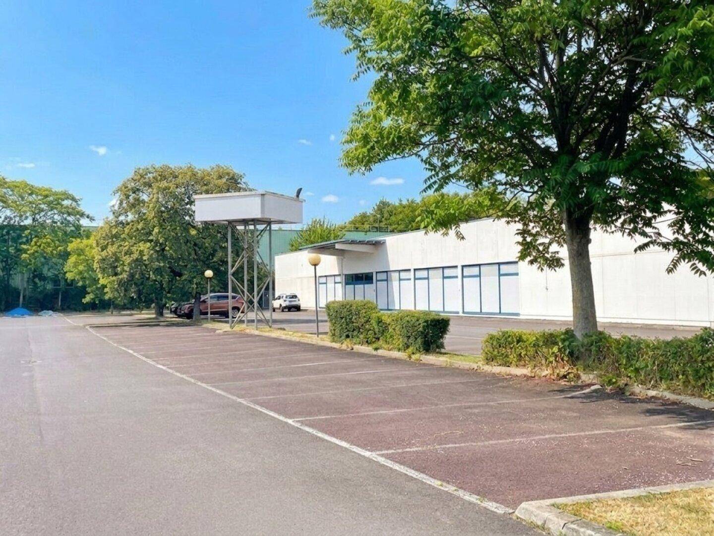 Außenansicht eines Gewerbegebäudes mit einem weitläufigen Parkplatz und grünen Bäumen unter klarem Himmel.