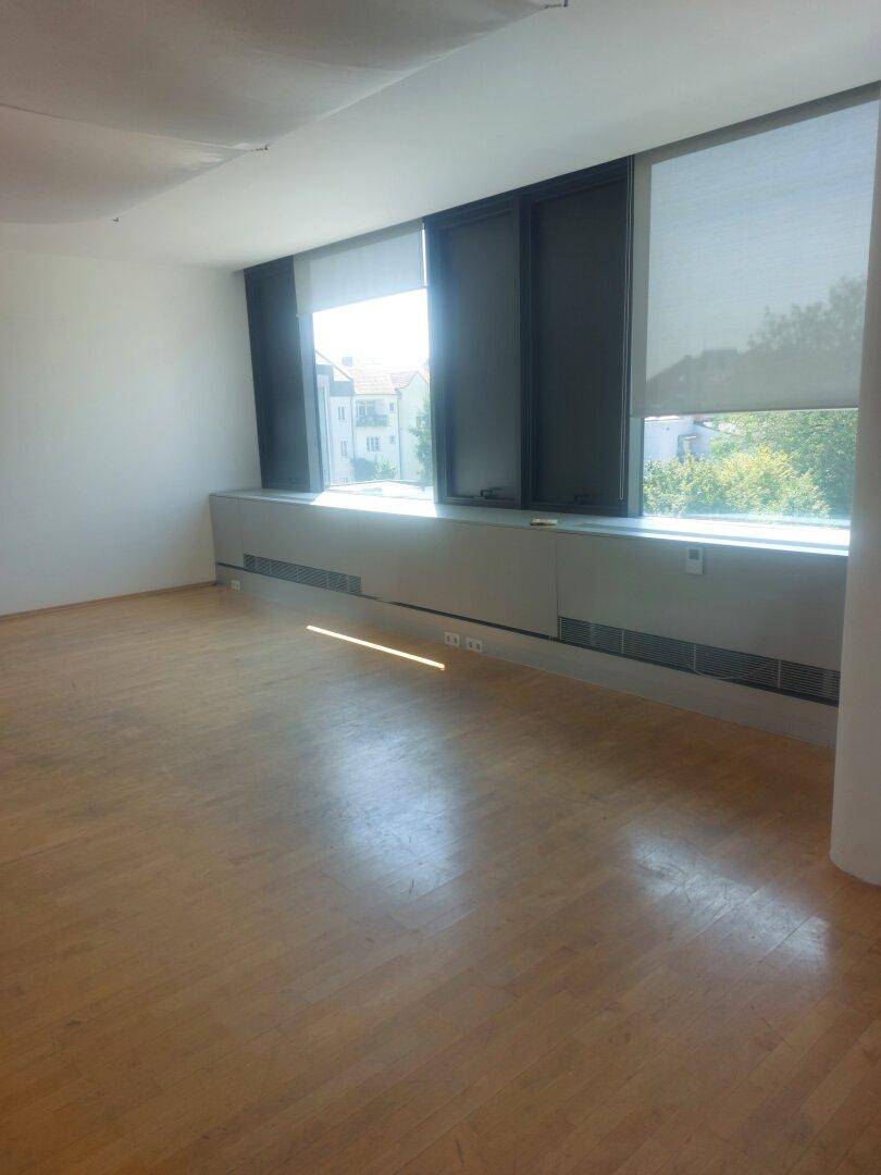 Heller, leerer Raum mit Holzboden und großen Fenstern, die viel Tageslicht hereinlassen.