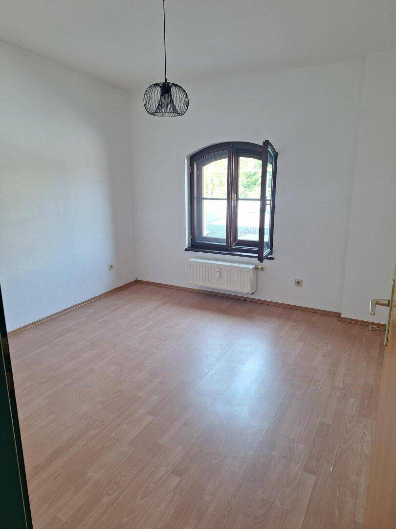 Helles Schlafzimmer mit Laminatboden, weiß gestrichenen Wänden und einem Fenster mit Blick ins Grüne.