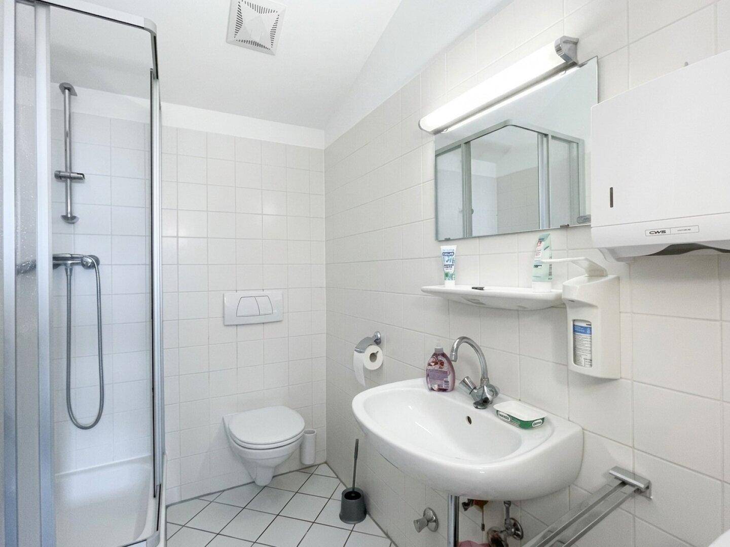 Funktionales Badezimmer mit Duschkabine, WC, Waschbecken und hellen Fliesen an Wänden und Boden.