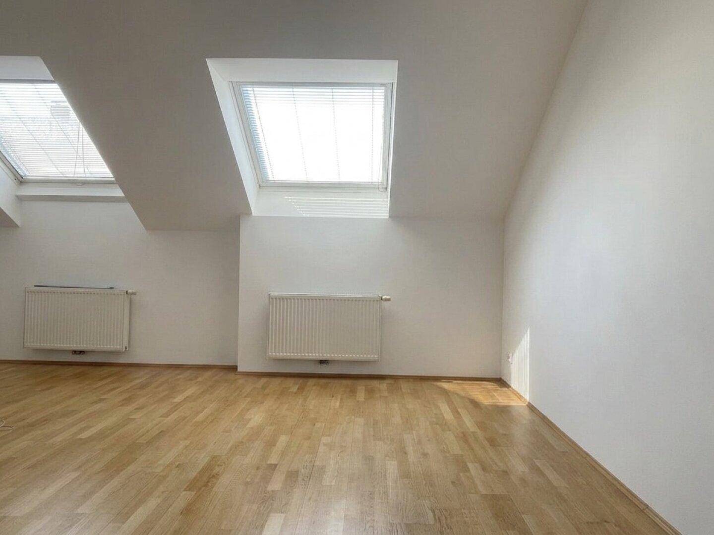 Leerer Raum im Dachgeschoss mit Holzboden, schrägen Wänden und zwei Dachfenstern.