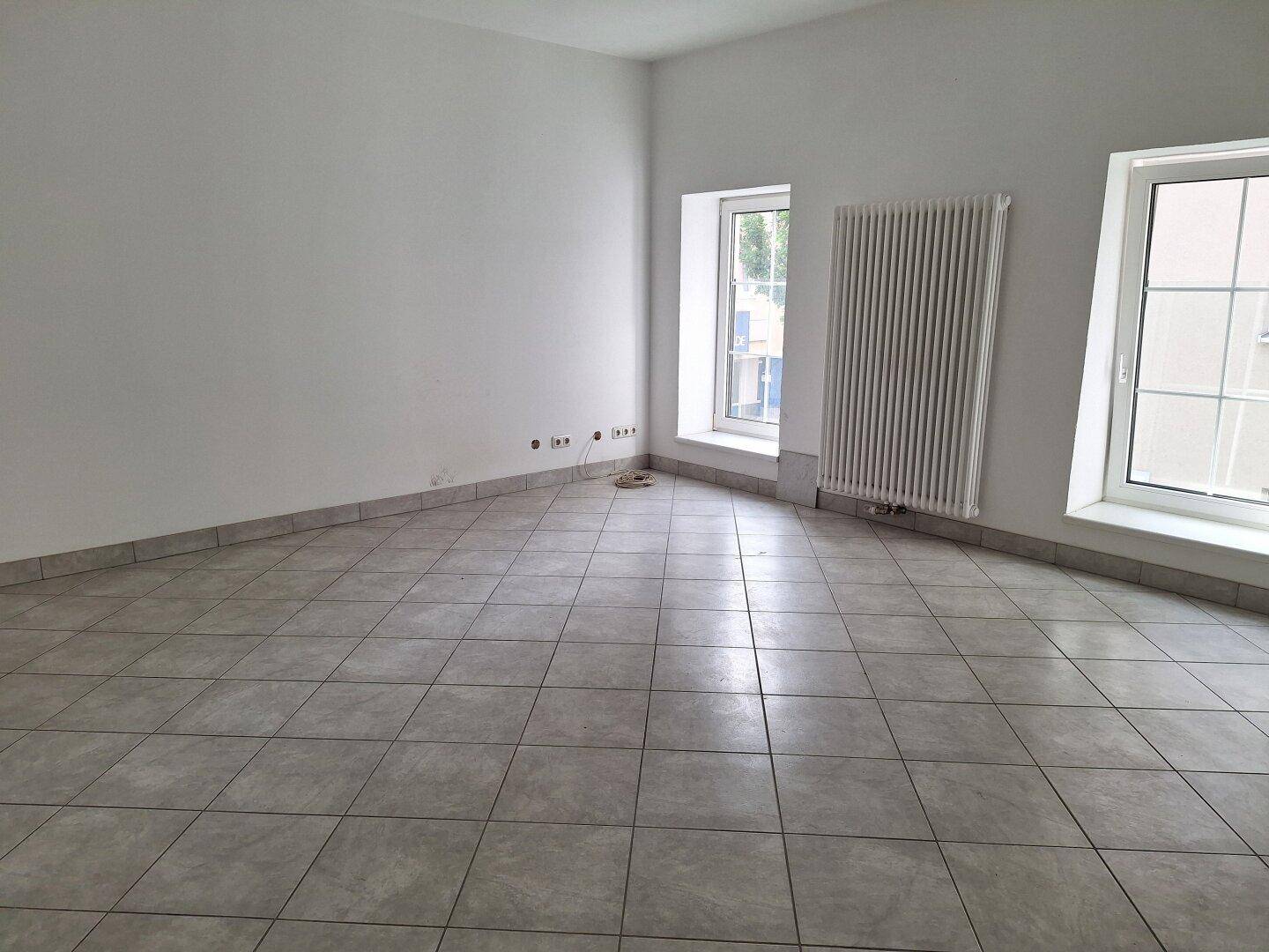 Heller, leerer Raum mit Fliesenboden, zwei Fenstern und einem großen Heizkörper an der Wand.