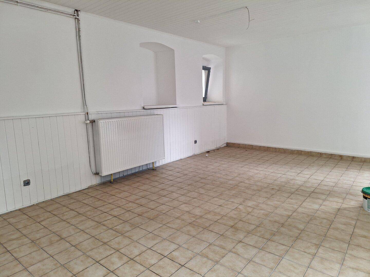 Leerer Raum mit hellen Fliesen, weiß getäfelten Wänden und einem Heizkörper unter einem kleinen Fenster.