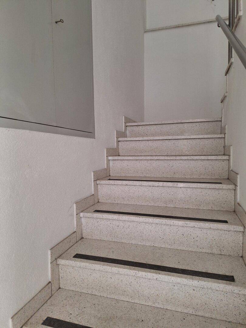 Helles Treppenhaus mit Terrazzo-Stufen, schwarzem Antirutschstreifen und Metallgeländer.