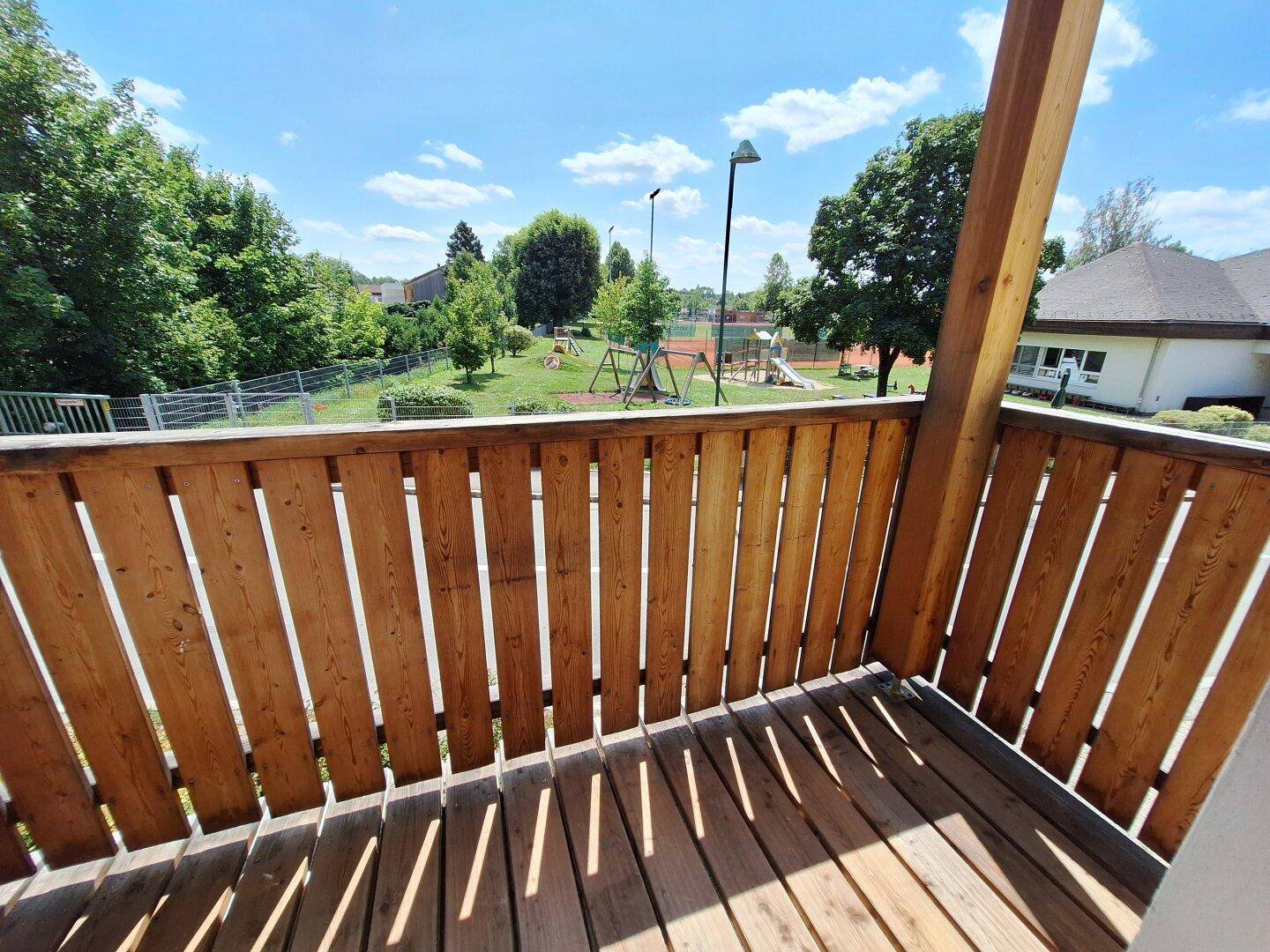 Holzbalkon mit Blick auf einen Spielplatz und grüne Umgebung.
